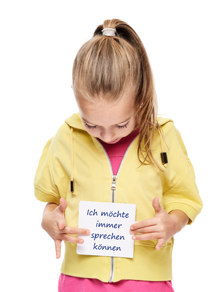 Ein kleines Mädchen mit langen blonden Haaren und gelber Sweetjacke steht mit gesenktem Kopf da. Sie schaut auf einen Zettel in ihren Händen auf dem steht: Ich möchte immer sprechen können