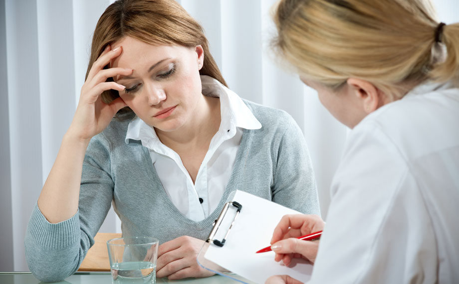 Eine junge langhaarige Frau sitzt mit gesenktem Blick und aufgestütztem Kopf am Tisch einer Therapeutin