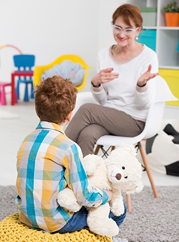 Eine Therapeutin spricht mit einem kleinen rothaarigen Jungen, der auf einem gelben Sitzkissen sitzt und einen großen Teddy im Arm hält.