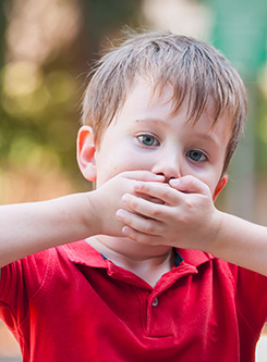 Kleiner Junge mit rotem Tshirt hält sich die Hände vor den Mund um zu zeigen, dass er nicht sprechen kann.