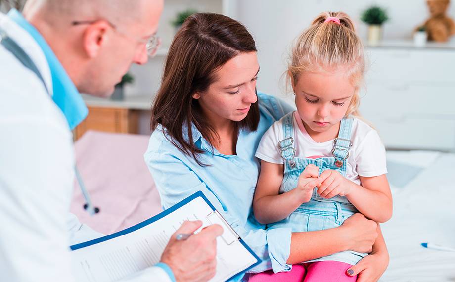 Ein kleines blondes Mädchen sitzt bei ihrer Mutter auf dem Schoss und scheut sich vor dem Blickkontakt mit dem Arzt