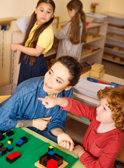 Kindergartenszene - Im Vordergrund beschäftigt sich eine Erzieherin mit einem kleinen rothaarigen Jungen, der mit dem Finger auf etwas zeigt. Im Hintergrund sieht 2 Mädchen spielen.